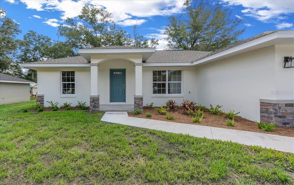 A single-story house with a white stucco exterior, teal front door, and stone-accented porch columns — 13413 SE 101ST TERRACE, BELLEVIEW, FL