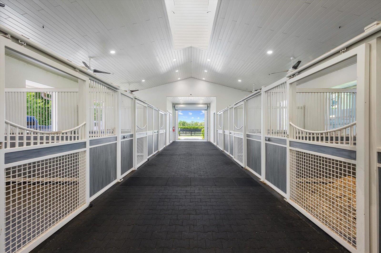 A bright horse stable aisle features white stalls with grey panels and black rubber flooring — 15450 Estancia Lane, Wellington FL 33414, WELLINGTON, FL