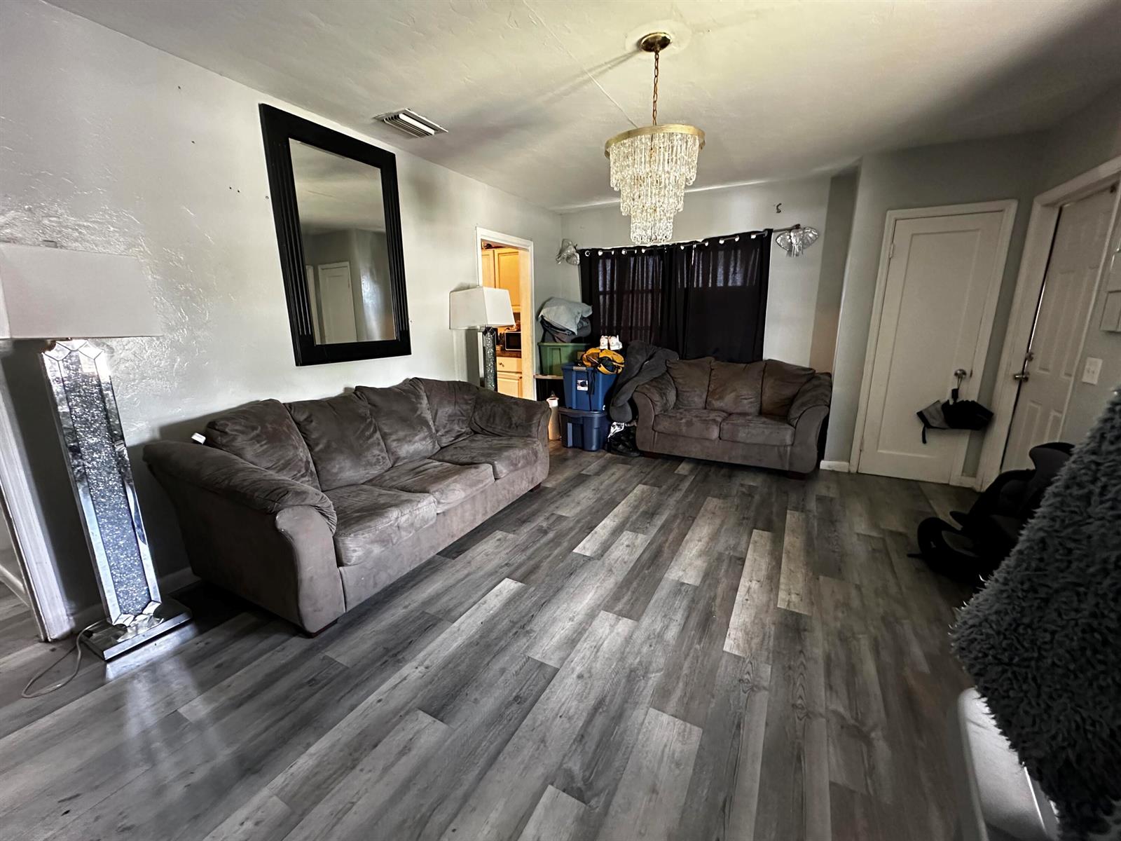 A living room features two brown sofas, a crystal chandelier, and gray wood-look flooring — 566 SE 7th Drive, Belle Glade FL 33430, BELLE GLADE, FL