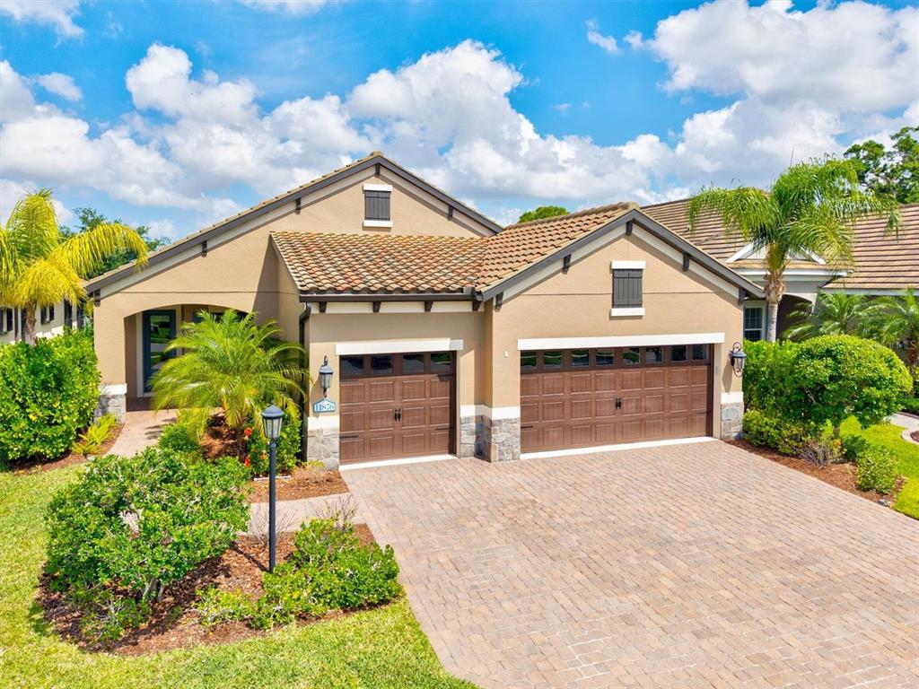 A tan single-story house features a paver driveway, two garage doors, and a tiled roof — 11876 HUNTERS CREEK ROAD, VENICE, FL