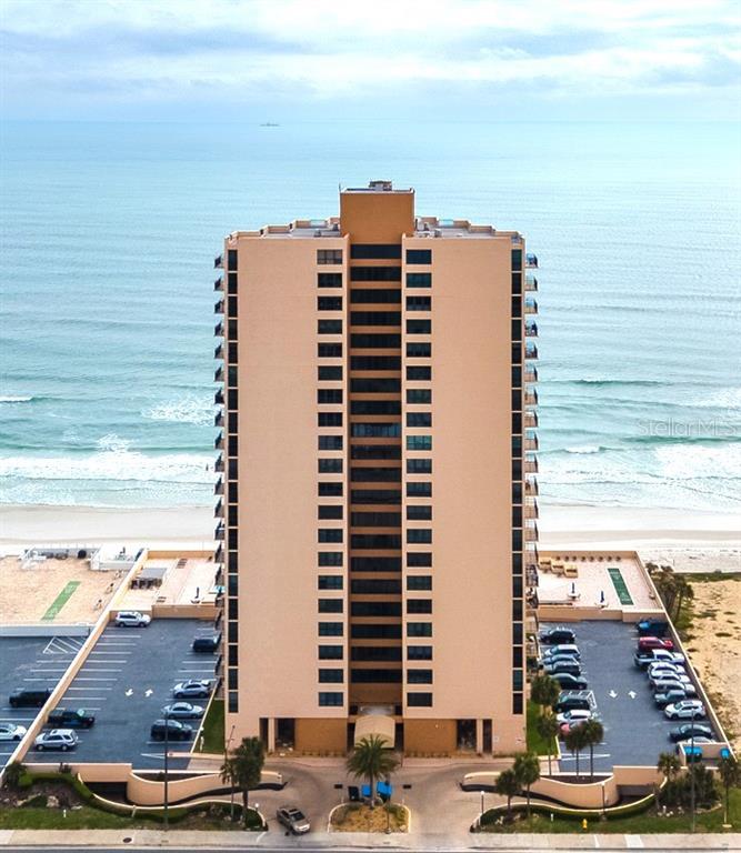 An aerial view of a tall, tan beachfront condominium building with parking lots on both sides — 3051 S. ATLANTIC AVENUE UNIT 1805, DAYTONA BEACH SHORES, FL
