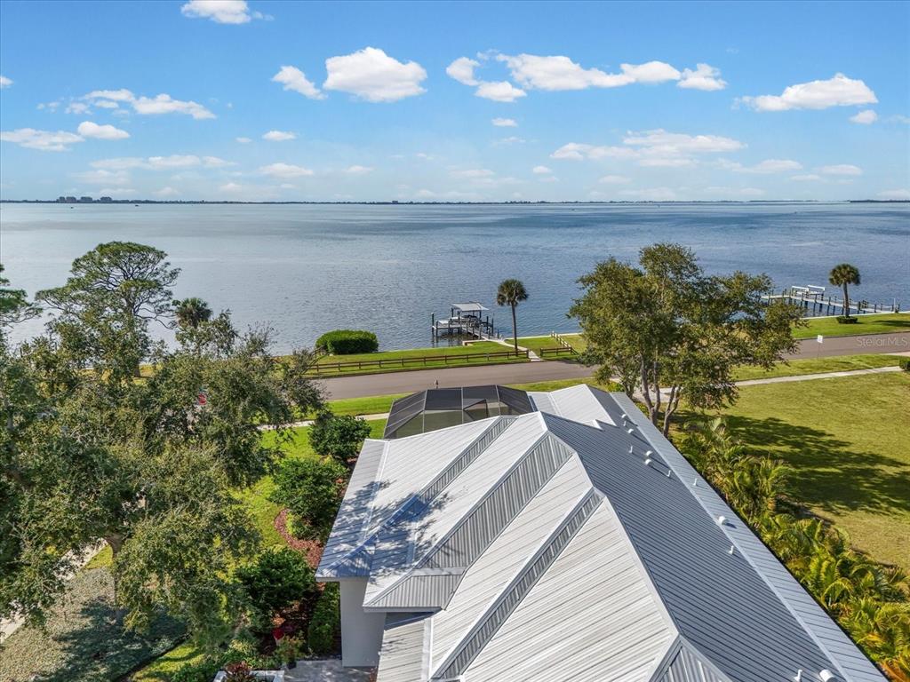 An aerial view shows a house with a metal roof overlooking a large lake — 7927 LONGBAY BOULEVARD, SARASOTA, FL