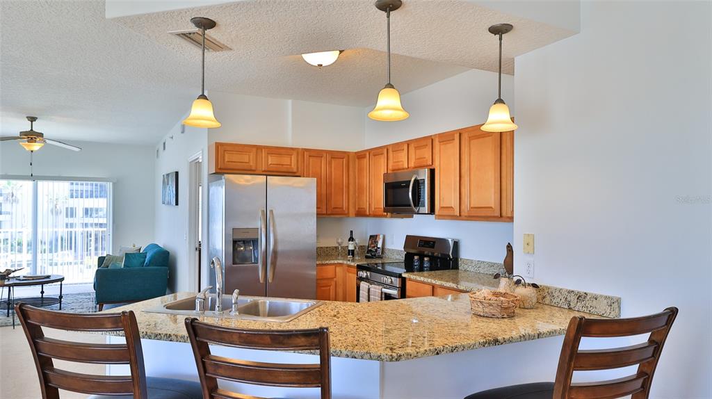A kitchen with wood cabinets, granite countertops, and stainless steel appliances overlooks a living area — 3756 S ATLANTIC AVENUE UNIT 201, DAYTONA BEACH, FL
