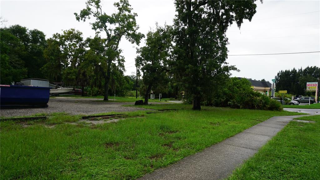 A gravel lot with a blue dumpster sits next to a sidewalk and mature trees — 7317 LITTLE ROAD, NEW PORT RICHEY, FL