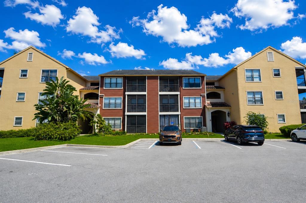 A three-story apartment building with tan and brown siding sits behind a paved parking lot — 11901 4TH STREET N UNIT 7204, ST PETERSBURG, FL