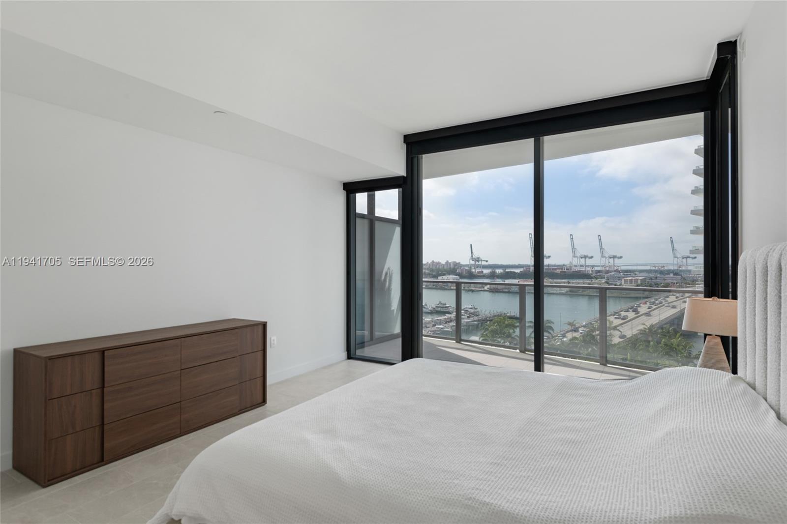 A bedroom features a bed, a wooden dresser, and floor-to-ceiling windows overlooking a harbor — 500 Alton Rd # 1204, Miami Beach FL 33139, MIAMI BEACH, FL