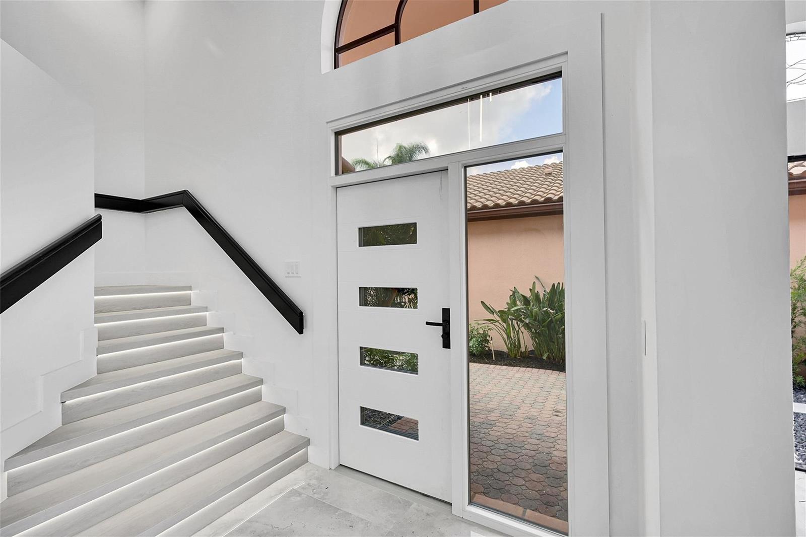 A modern entryway features a white front door with glass panels and an illuminated staircase — 6488 Via Rosa, Boca Raton FL 33433, BOCA RATON, FL