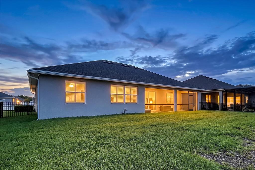 The back exterior of a house with a screened-in lanai and a grassy backyard at dusk — 6104 HIGHLANDS GRACE BOULEVARD, LAKELAND, FL