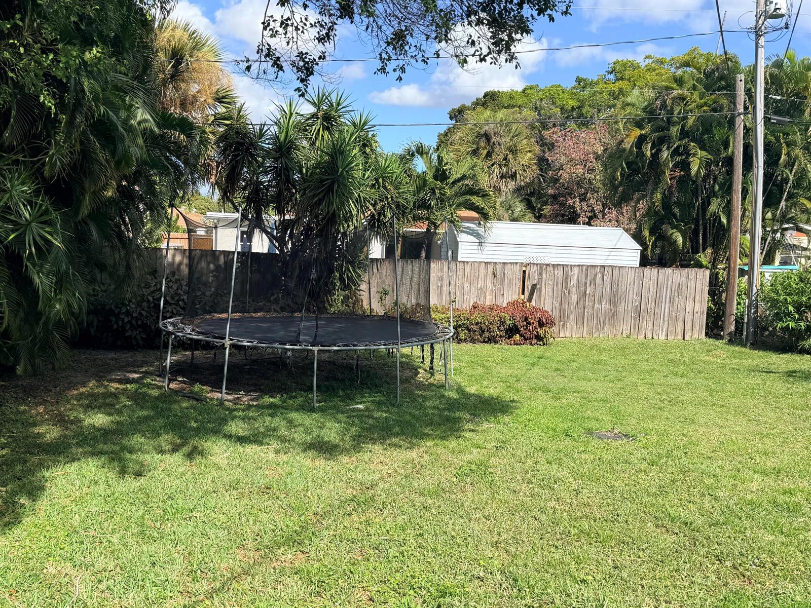 A backyard features a trampoline, a wooden fence, and various trees under a blue sky — 937 Upland Road, West Palm Beach FL 33401, WEST PALM BEACH, FL