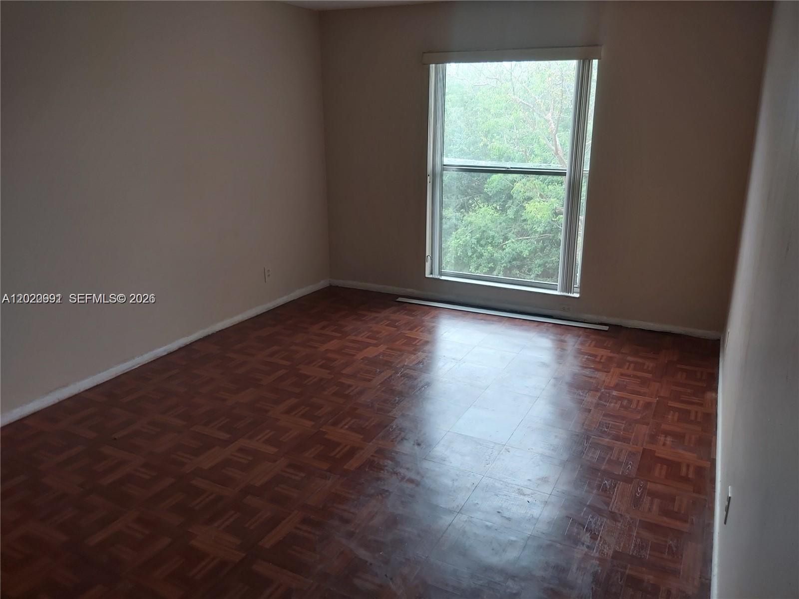 An empty room features parquet wood flooring, beige walls, and a large window overlooking trees — TAMARAC, FL