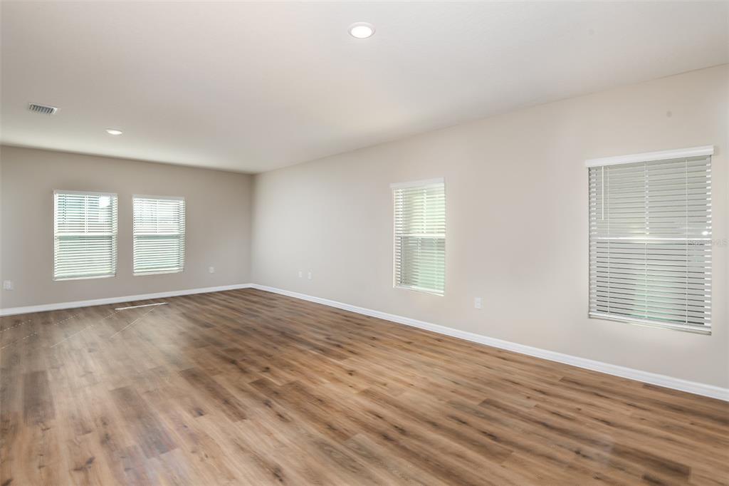 An empty living room with light-colored walls, wood-look flooring, and several windows with blinds — 33669 NOBLE CREEK PLACE, DADE CITY, FL
