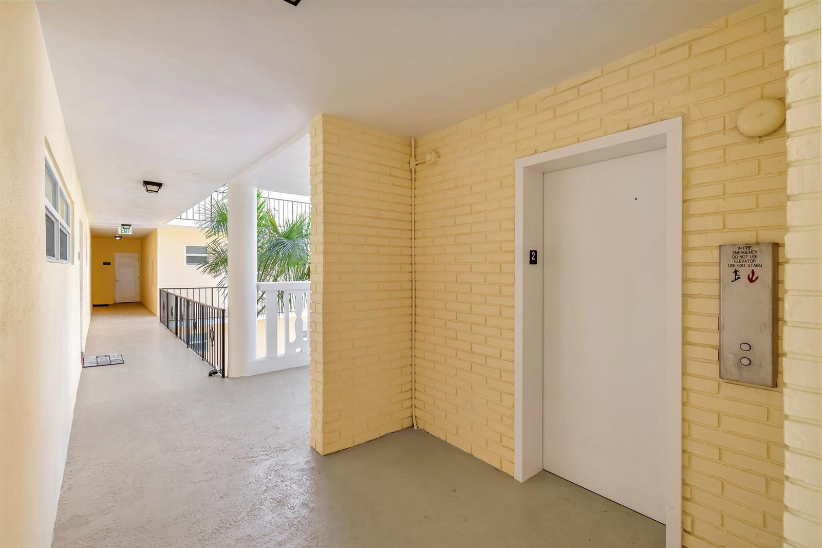 An outdoor apartment hallway features a yellow brick wall, an elevator, and a metal railing — 17 Colonial Club Drive # 204, Boynton Beach FL 33435, BOYNTON BEACH, FL