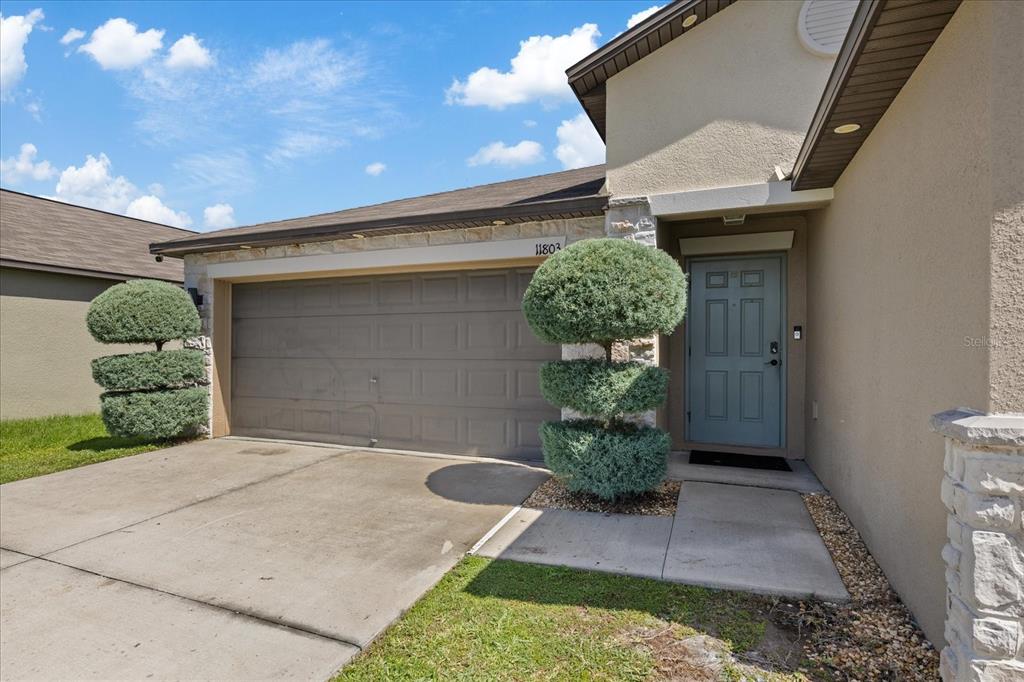 A tan house exterior features a two-car garage, a blue front door, and manicured shrubs — 11803 BLACKEYED SUSAN DRIVE, RIVERVIEW, FL