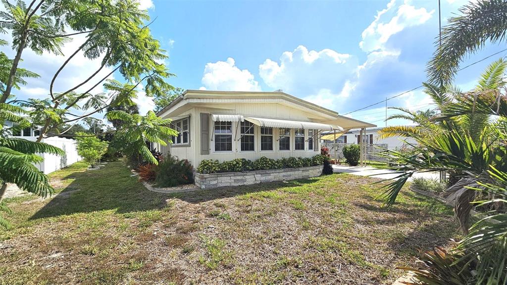 A light-colored mobile home features a stone-veneer base, metal awnings, and surrounding tropical landscaping — 1240 SEAGULL DRIVE, ENGLEWOOD, FL
