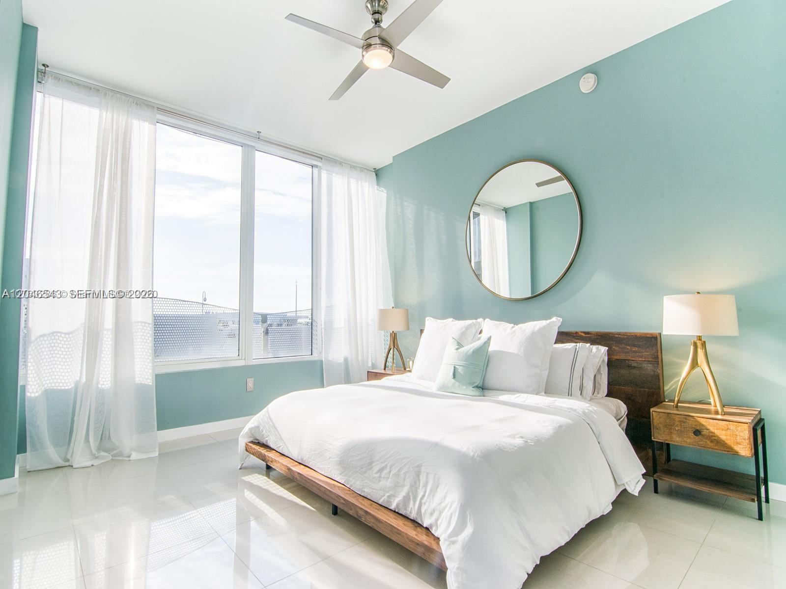 A bedroom features a platform bed, a round mirror, and large windows with white curtains — 2100 Park Ave # 406, Miami Beach FL 33139, MIAMI BEACH, FL