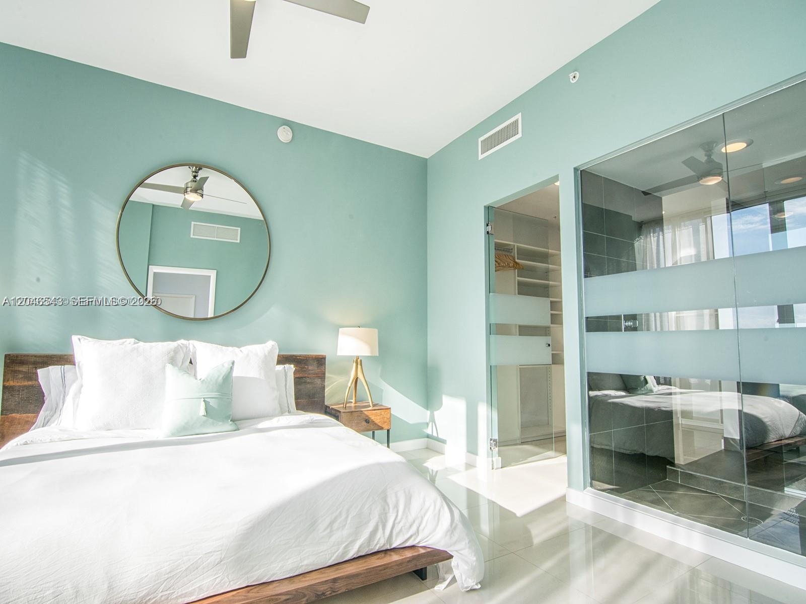 A bedroom features light blue walls, a wooden bed frame, a round mirror, and glass-enclosed areas — 2100 Park Ave # 406, Miami Beach FL 33139, MIAMI BEACH, FL
