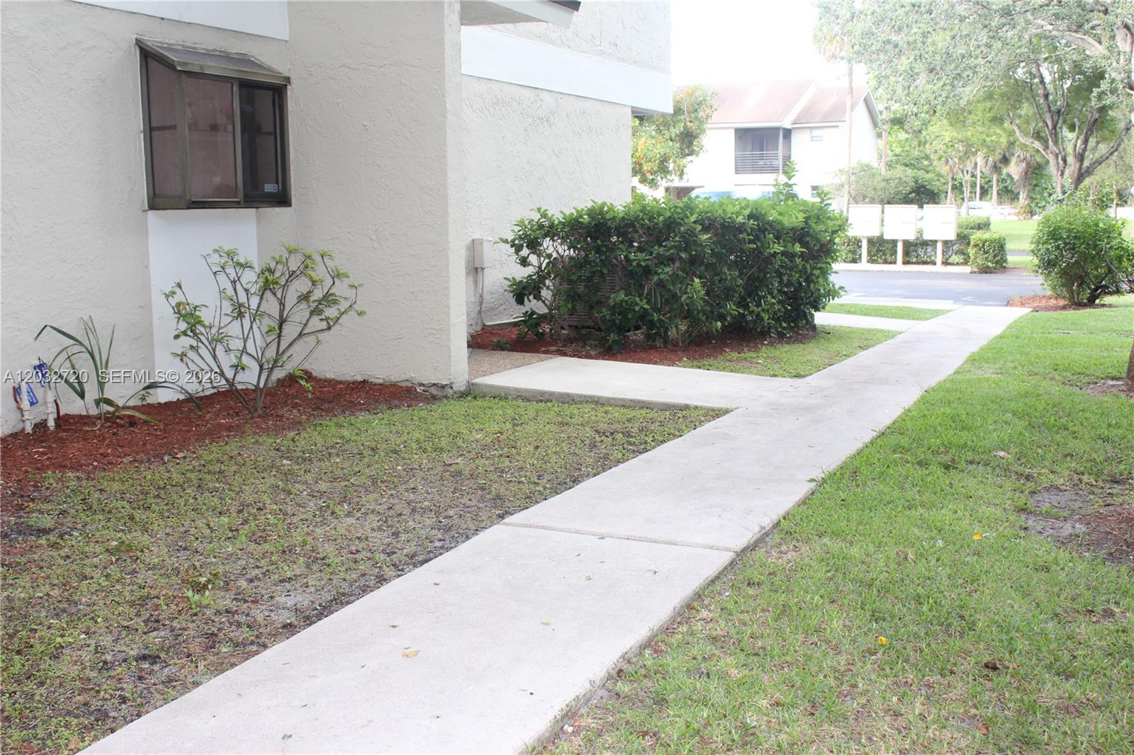 A concrete walkway leads to the side entrance of a beige stucco building — 110 Gardens Dr # 103, Pompano Beach FL 33069, POMPANO BEACH, FL