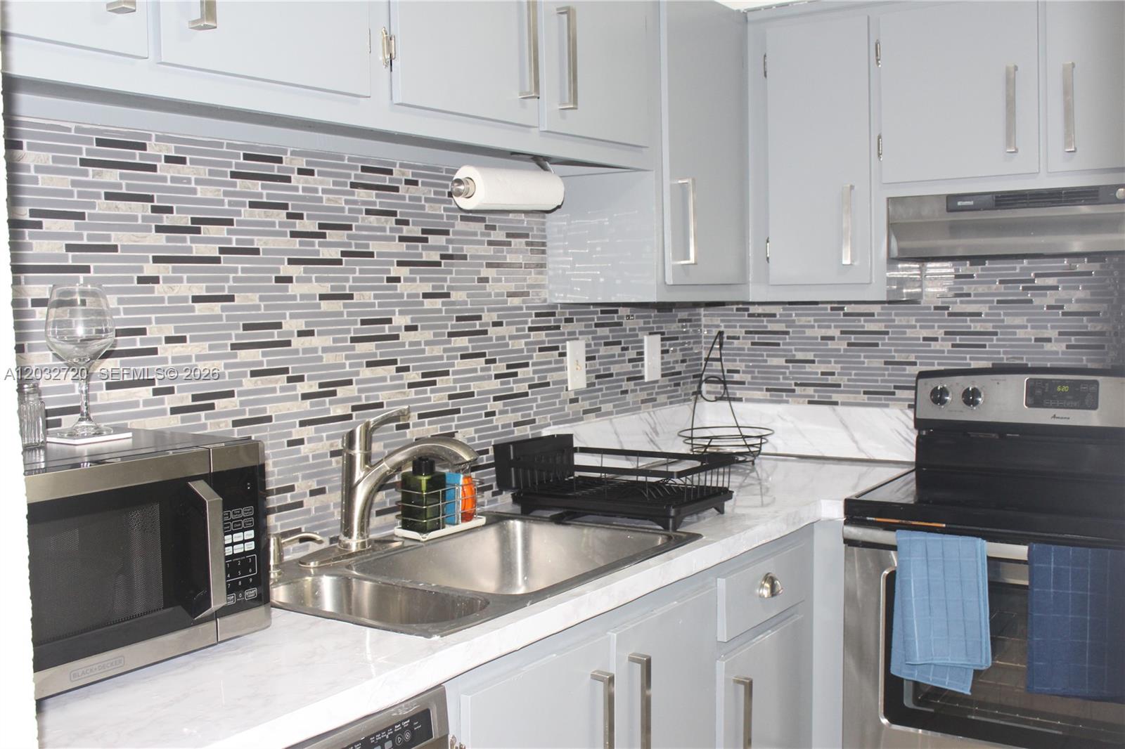 A kitchen features gray cabinets, a mosaic tile backsplash, stainless steel appliances, and marble countertops — 110 Gardens Dr # 103, Pompano Beach FL 33069, POMPANO BEACH, FL