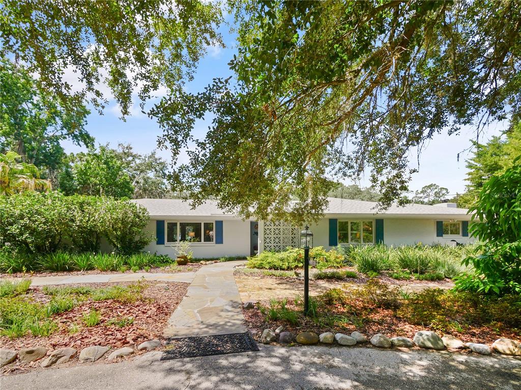 A single-story white house with blue shutters sits behind a stone walkway and large trees — 5701 CROSS FOX LANE, OVIEDO, FL