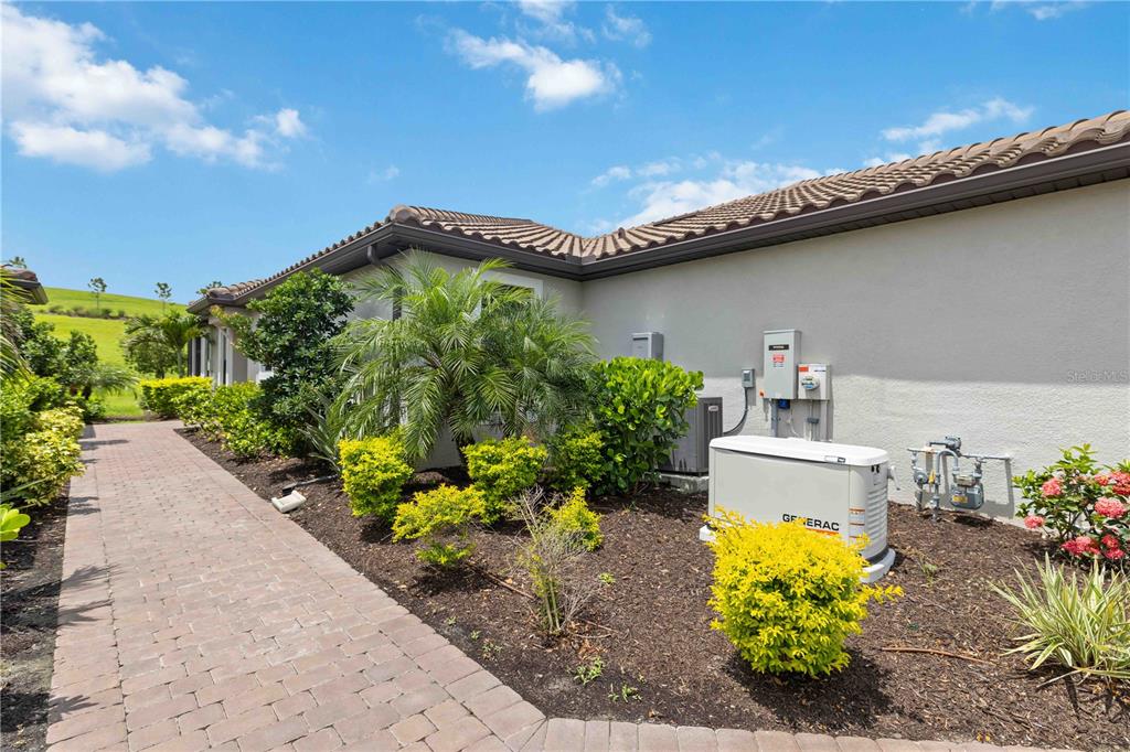A side view of a house exterior featuring a brick walkway, landscaping, and a generator — 6242 GRANDVIEW HILL COURT, BRADENTON, FL