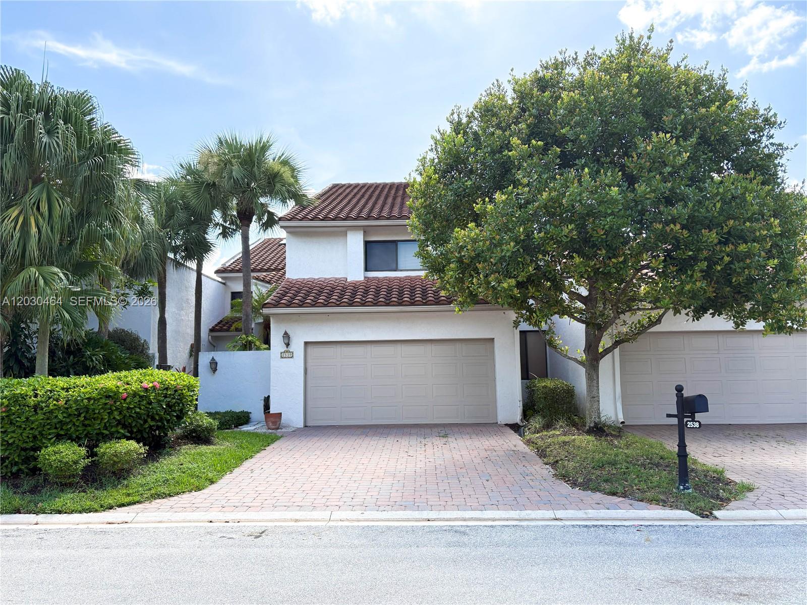 A two-story house features a beige garage, a brick driveway, and a large tree — 2530 Windsor Way Ct, Wellington FL 33414, WELLINGTON, FL