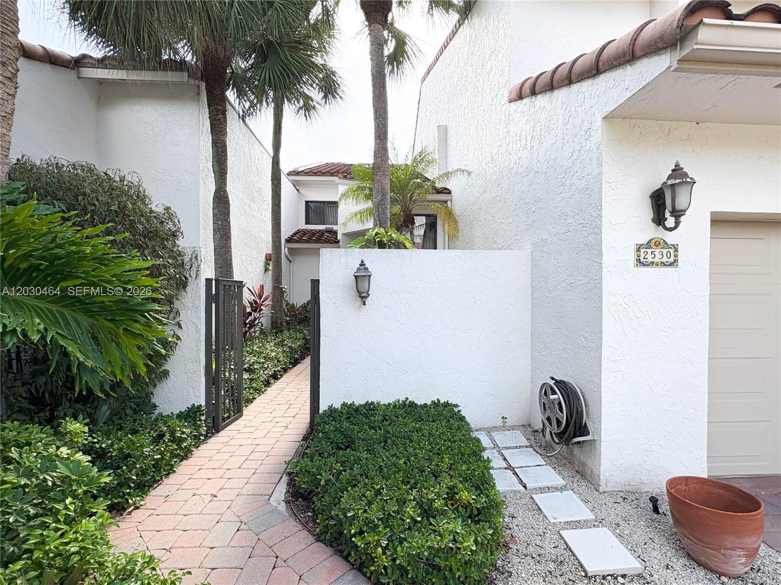 A brick walkway leads through a gate toward a white stucco house with palm trees — 2530 Windsor Way Ct, Wellington FL 33414, WELLINGTON, FL