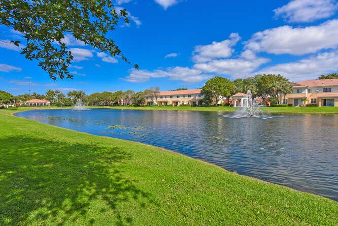 A pond with two fountains sits in front of apartment buildings under a blue sky — 7401 Sarentino Lane # 211, Boynton Beach FL 33437, BOYNTON BEACH, FL