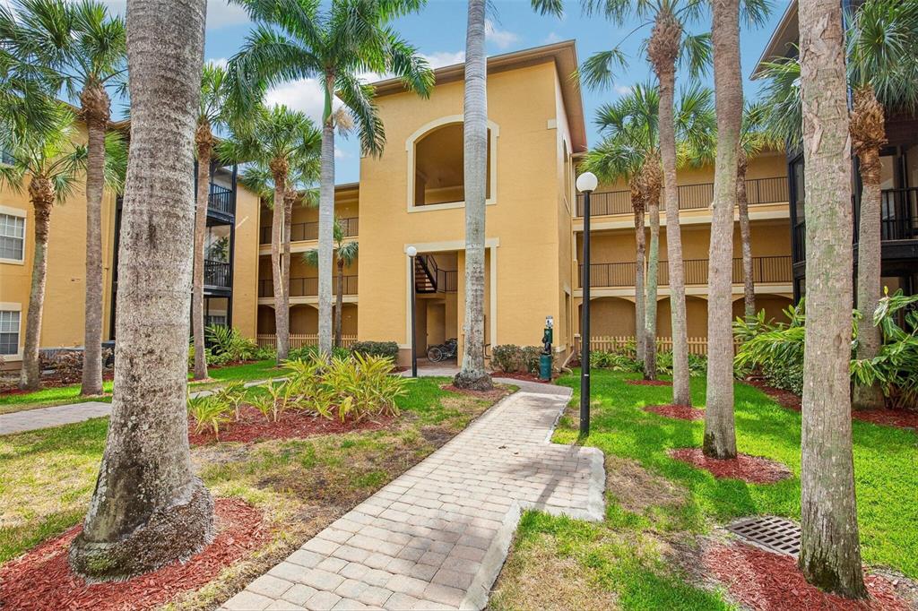 A paved walkway leads toward the entrance of a multi-story yellow apartment building with palms — 4333 BAYSIDE VILLAGE DRIVE UNIT 322, TAMPA, FL