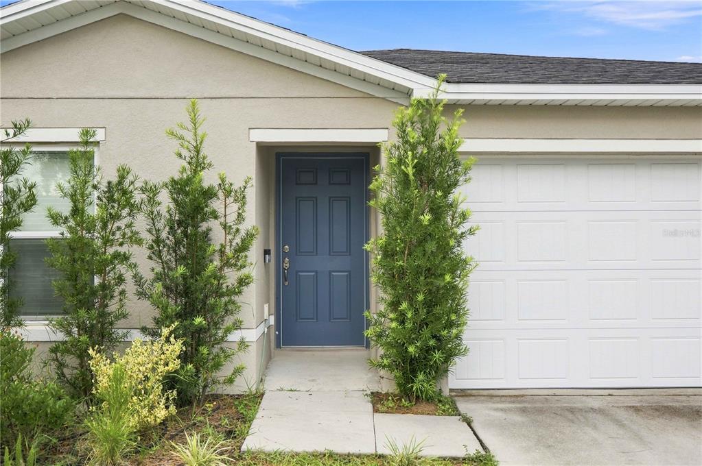 A blue front door is centered between two tall evergreen shrubs and a white garage — 1876 IOWA DRIVE, SUMTERVILLE, FL