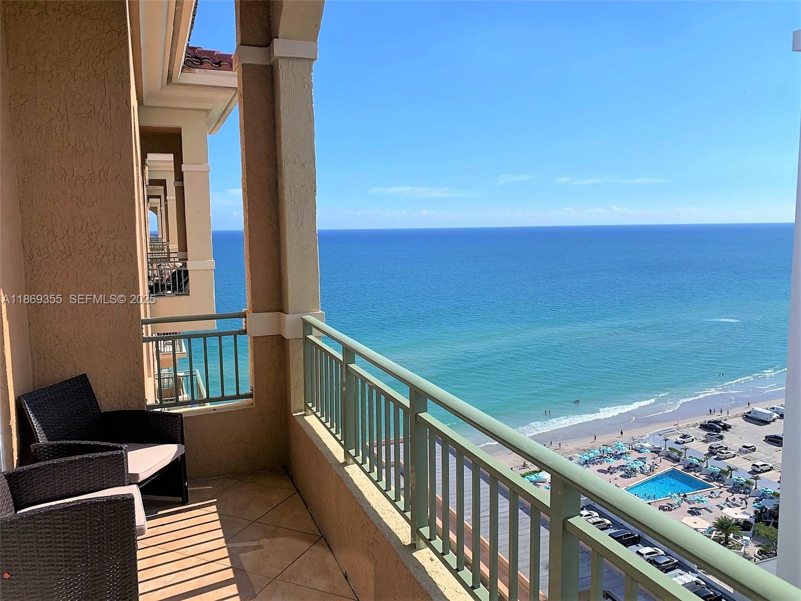 A balcony with wicker chairs overlooks the ocean, a beach, and a swimming pool below — 2080 S OCEAN DRIVE, Hallandale Beach FL 33009, HALLANDALE BEACH, FL