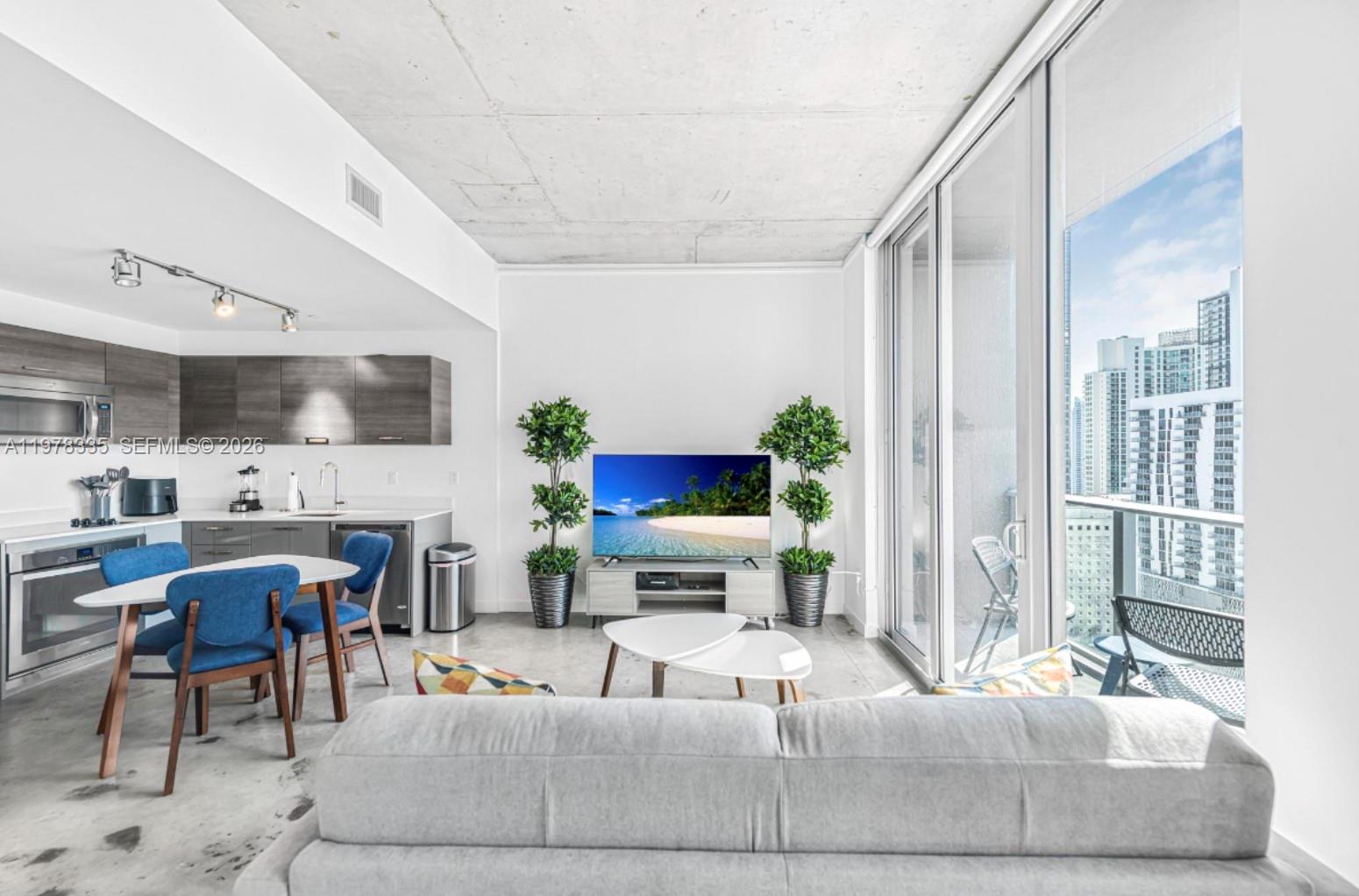 A modern living room and kitchen area with concrete ceilings and a balcony overlooking city buildings — 151 SE 1st St # 2409, Miami FL 33131, MIAMI, FL
