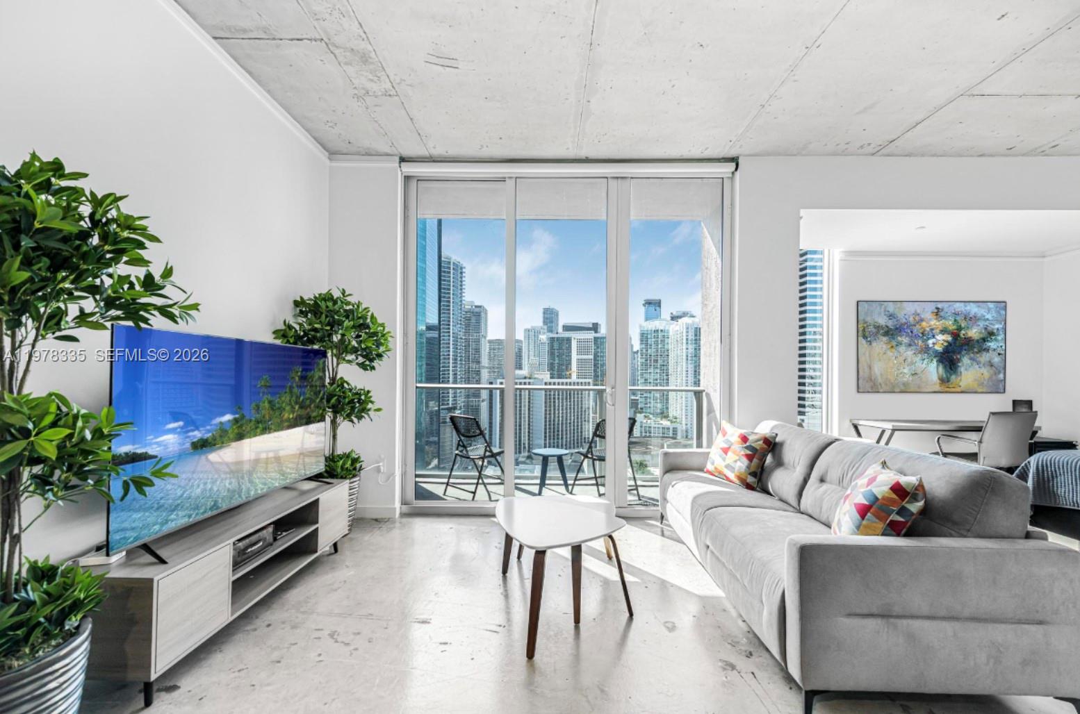 A modern living room features a grey sofa, television, and floor-to-ceiling windows with city views — 151 SE 1st St # 2409, Miami FL 33131, MIAMI, FL