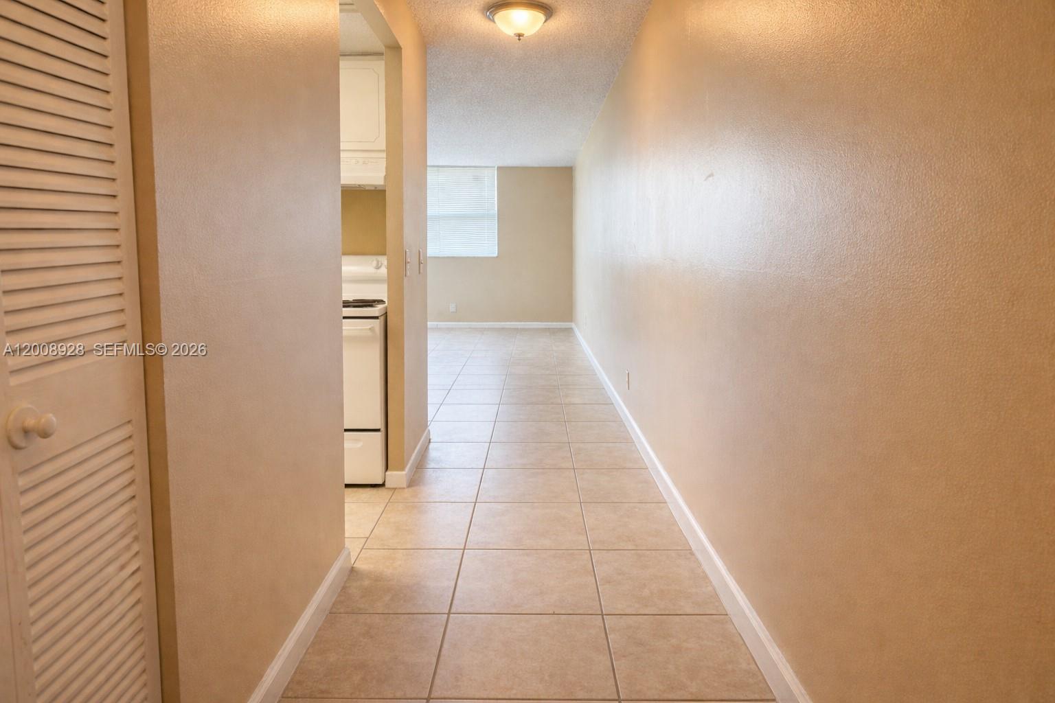 A hallway with beige tile flooring leads toward a kitchen and a windowed living area — 470 Executive Center Dr # 2D, West Palm Beach FL 33401, WEST PALM BEACH, FL