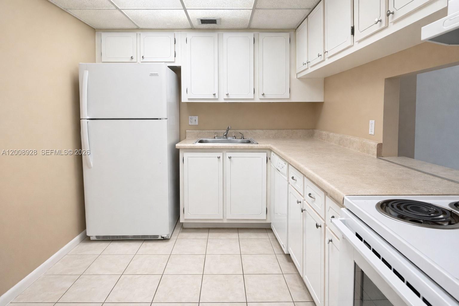 A kitchen features white cabinets, a white refrigerator, a sink, and beige countertops — 470 Executive Center Dr # 2D, West Palm Beach FL 33401, WEST PALM BEACH, FL