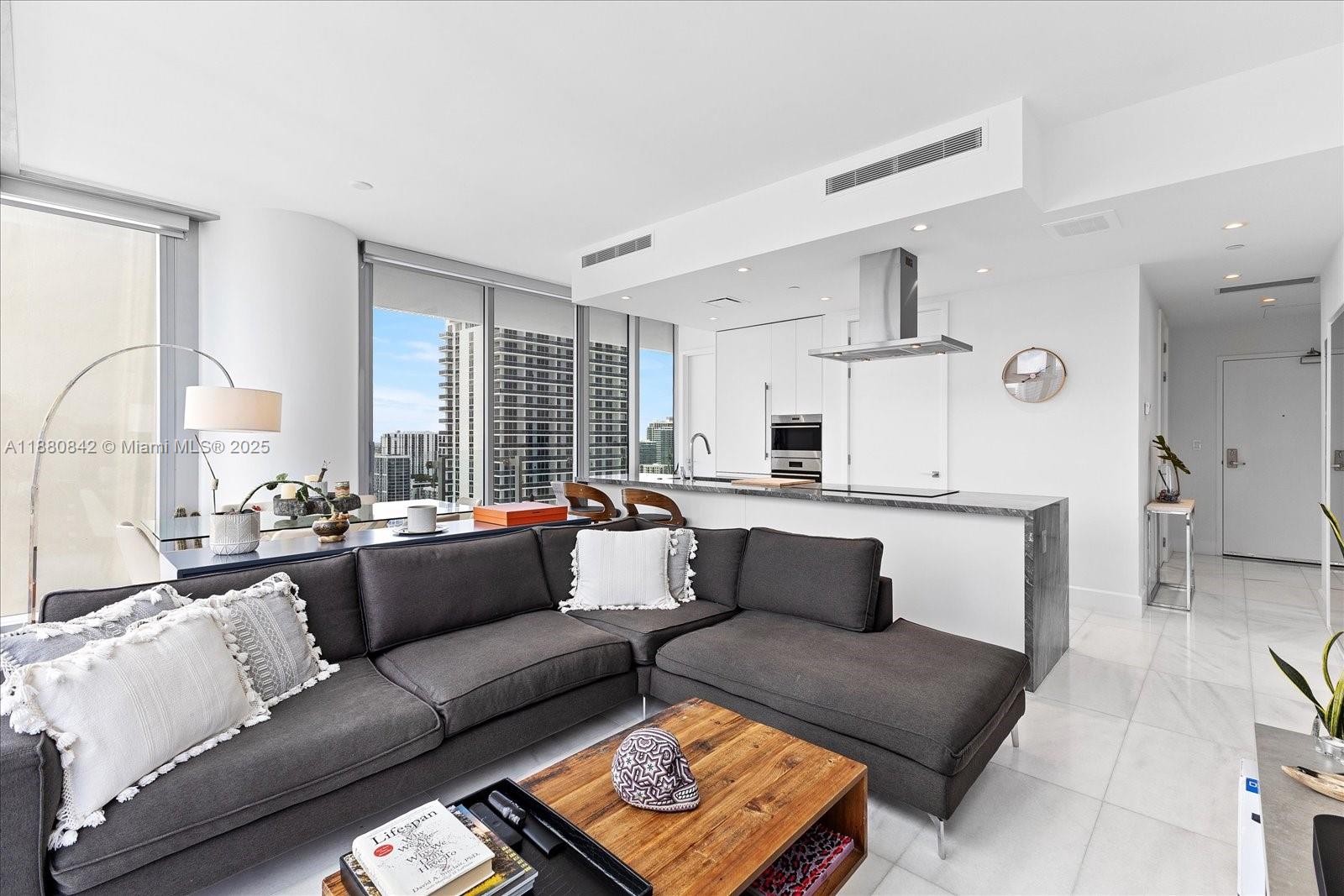 A modern living room and kitchen area with a dark sectional sofa and marble floors — 700 NE 26th Ter # 2605, Miami FL 33137, MIAMI, FL