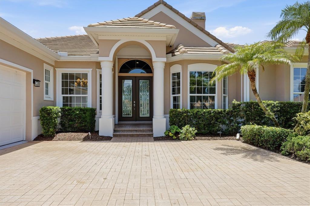 A tan house exterior features a paved driveway, arched entryway, and double glass front doors — 8160 COLLINGWOOD COURT, UNIVERSITY PARK, FL