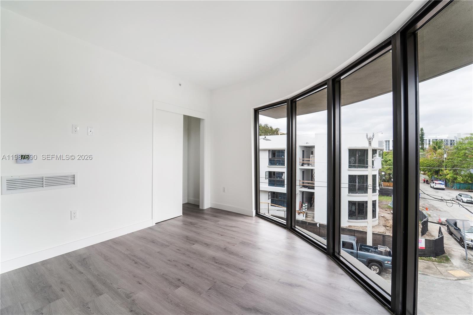 An empty room features white walls, dark wood-look flooring, and floor-to-ceiling windows overlooking construction — 1165 NW 8th Ave # 104, Miami FL 33136, MIAMI, FL