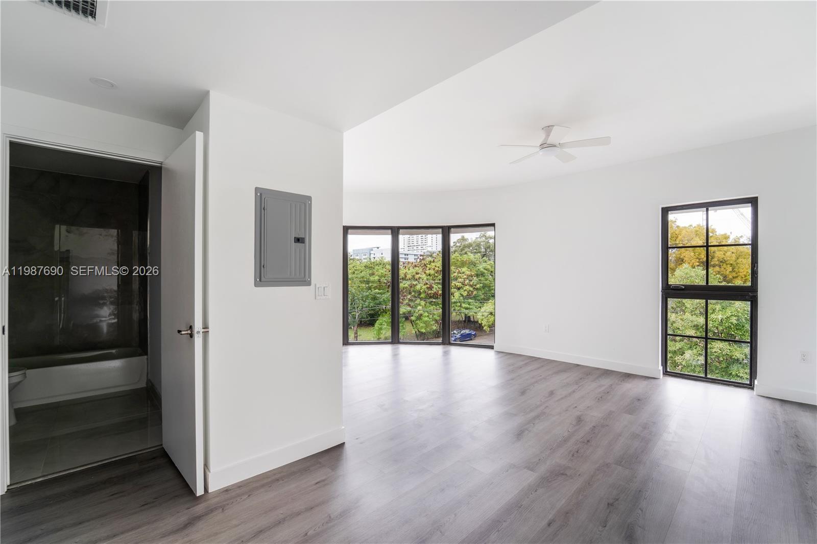 A bright room features gray wood-look flooring, a ceiling fan, and large windows overlooking trees — 1165 NW 8th Ave # 104, Miami FL 33136, MIAMI, FL