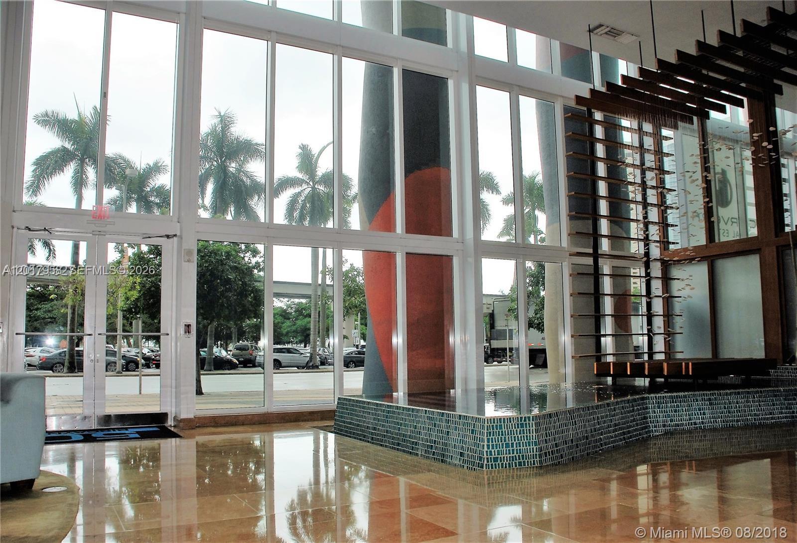 A modern lobby features floor-to-ceiling windows, a tiled water feature, and a large decorative column — 50 Biscayne Blvd # 1801, Miami FL 33132, MIAMI, FL