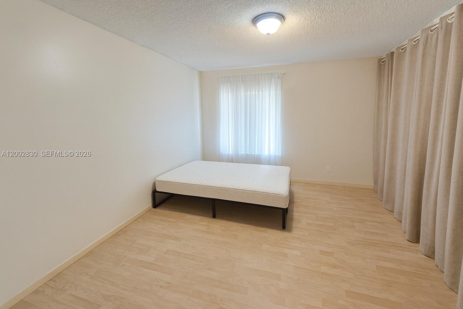 A bedroom features light wood-look flooring, white walls, a mattress on a frame, and curtains — 9999 Summerbreeze Dr # 216, Sunrise FL 33322, SUNRISE, FL