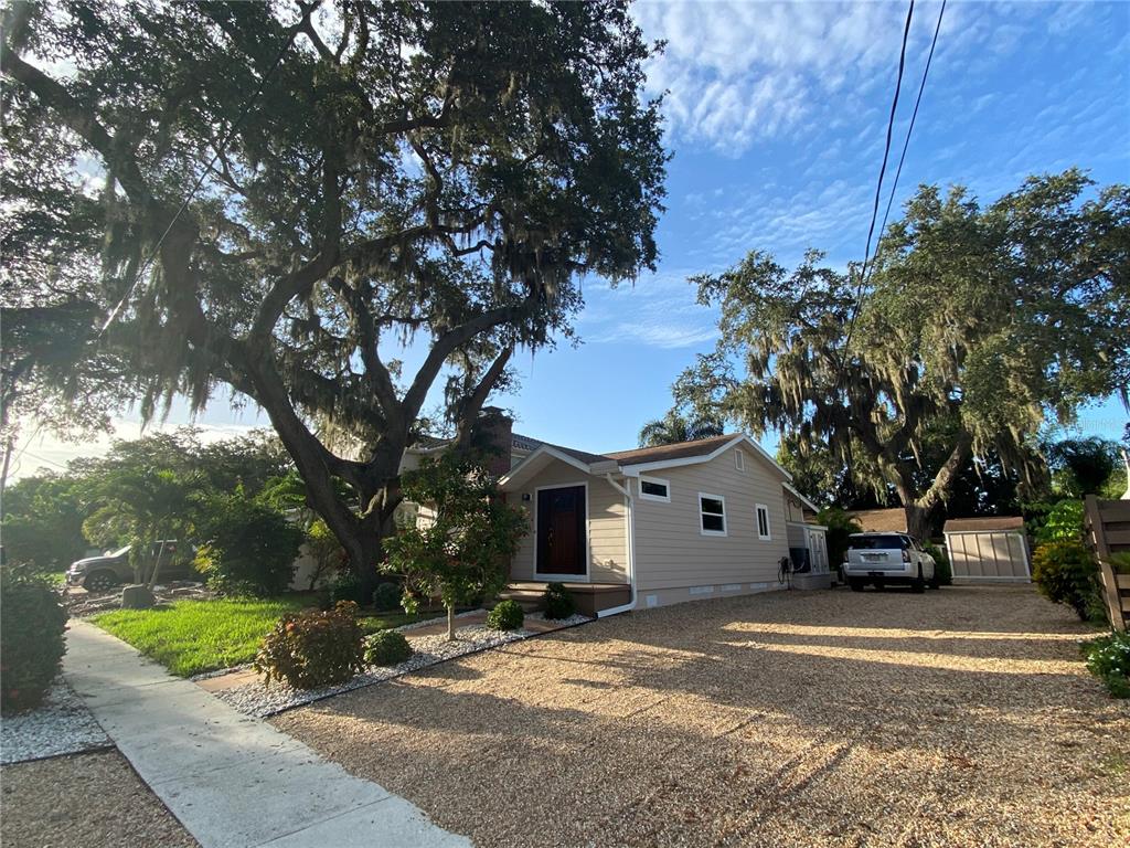 A tan-sided house with a gravel driveway sits under large oak trees on a sunny day — 1832 WORRINGTON STREET, SARASOTA, FL