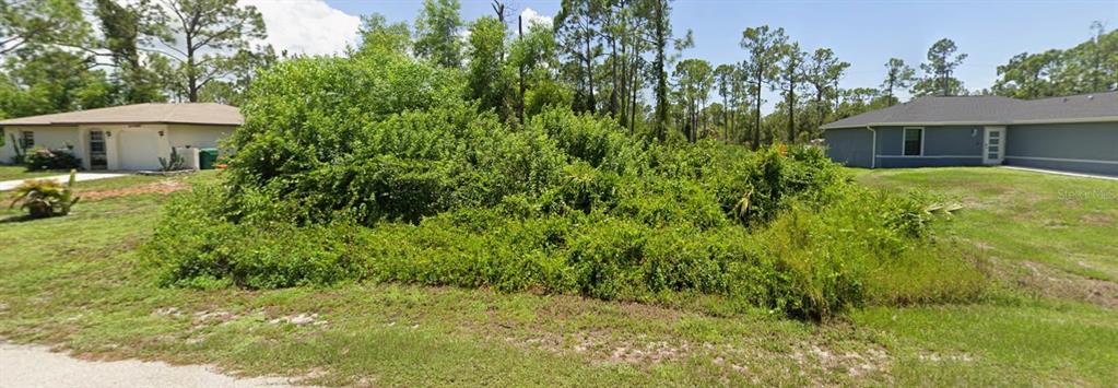 A vacant, overgrown lot sits between two suburban houses under a clear blue sky — 27077 N TWIN LAKES DRIVE, PUNTA GORDA, FL