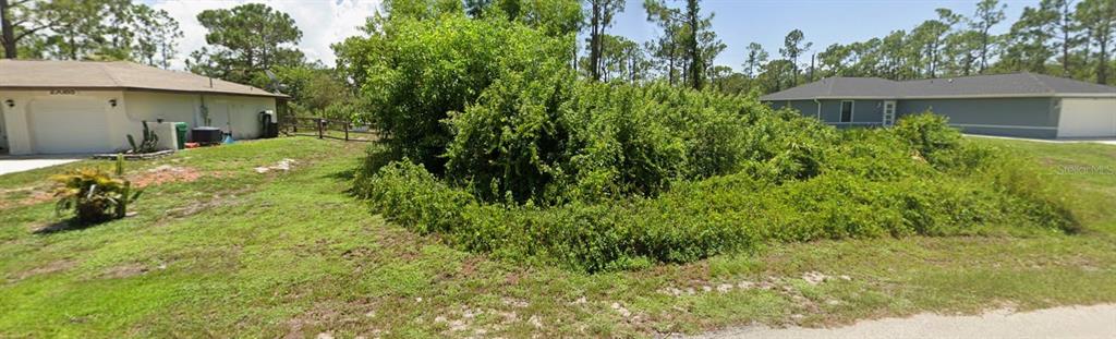 A vacant, overgrown lot sits between two suburban houses under a clear blue sky — 27077 N TWIN LAKES DRIVE, PUNTA GORDA, FL