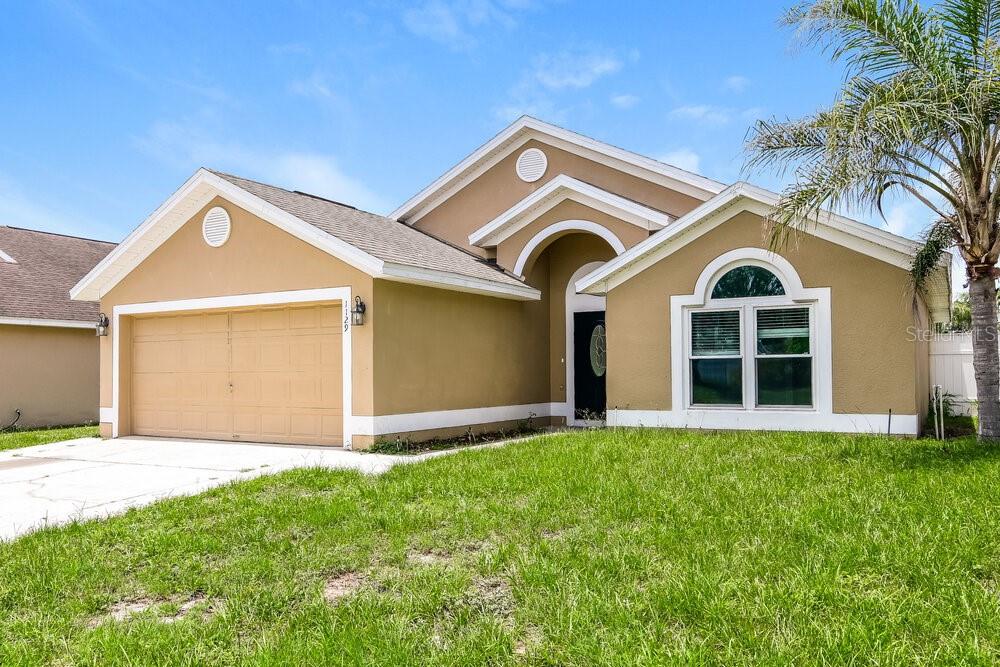 A tan, single-story suburban house with a two-car garage and a front-facing arched window — 1129 PINEDALE DRIVE, DAVENPORT, FL