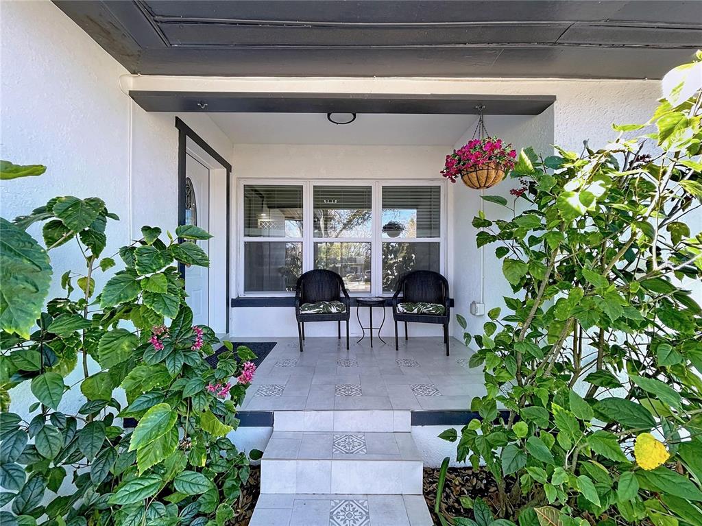A covered front porch features tiled steps, two wicker chairs, and surrounding green foliage — 1468 S PRESCOTT AVENUE, CLEARWATER, FL