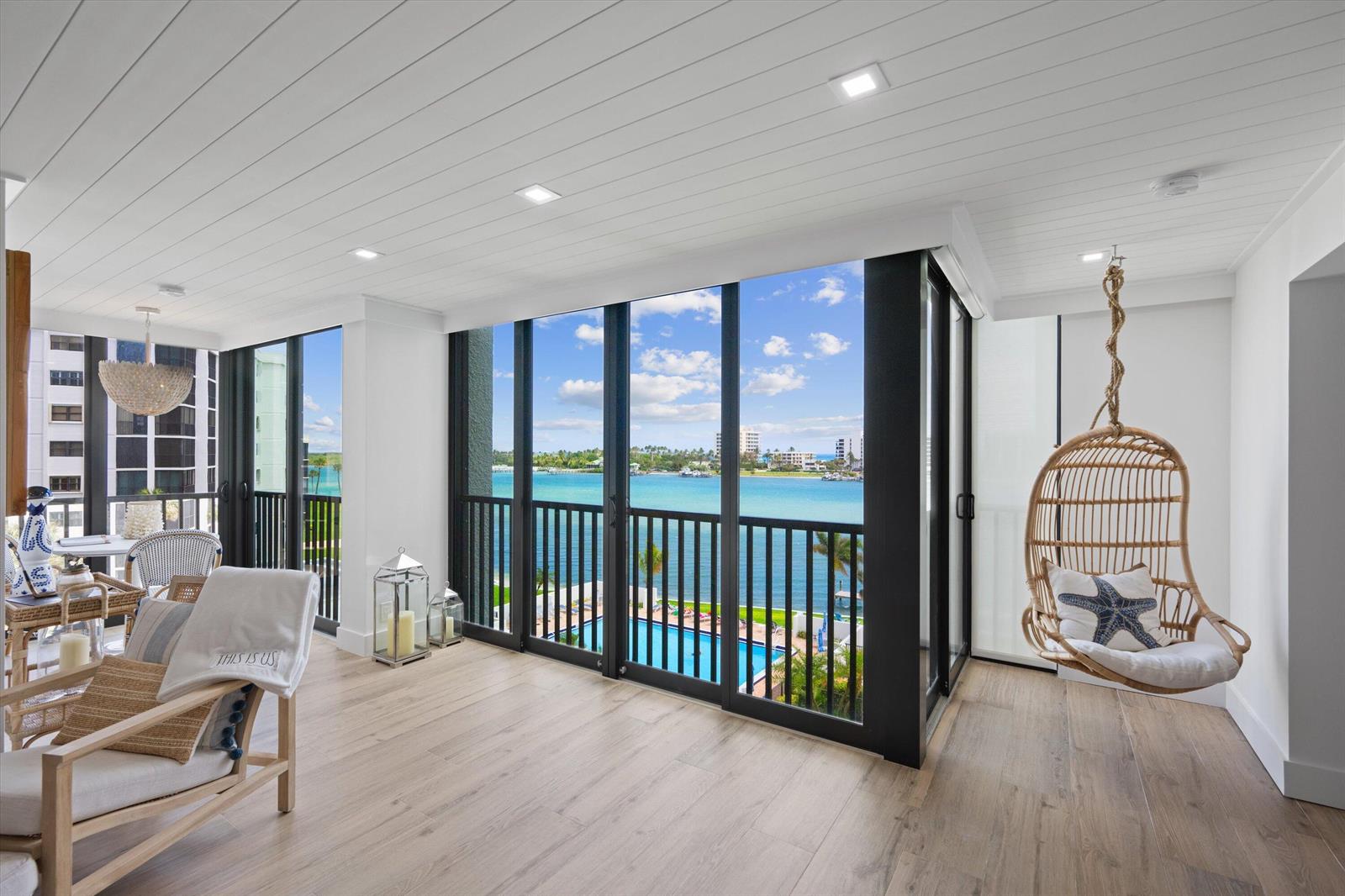 A bright sunroom features light wood floors, a hanging rattan chair, and floor-to-ceiling water views — 19800 S Sandpointe Bay Drive # 510, Tequesta FL 33469, TEQUESTA, FL