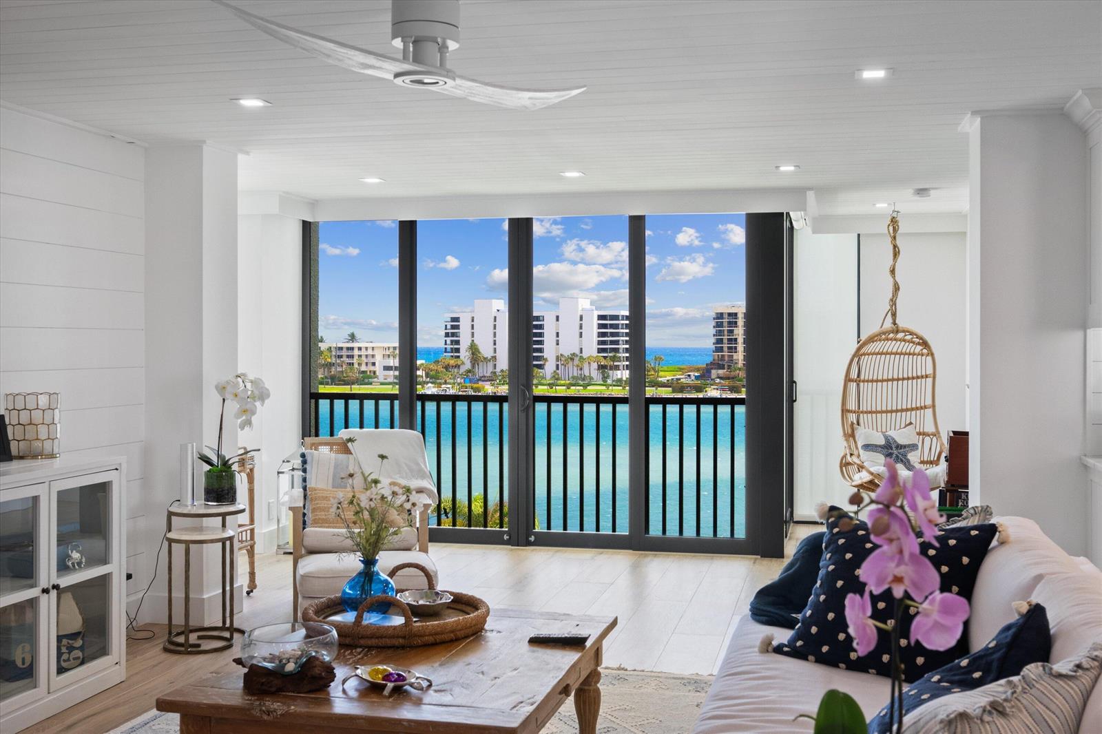 A bright living room features a wooden coffee table, hanging chair, and floor-to-ceiling water views — 19800 S Sandpointe Bay Drive # 510, Tequesta FL 33469, TEQUESTA, FL