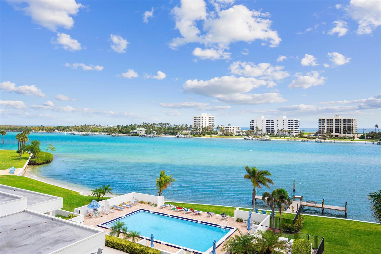 An elevated view of a swimming pool overlooking a turquoise bay with distant apartment buildings — 19800 S Sandpointe Bay Drive # 510, Tequesta FL 33469, TEQUESTA, FL