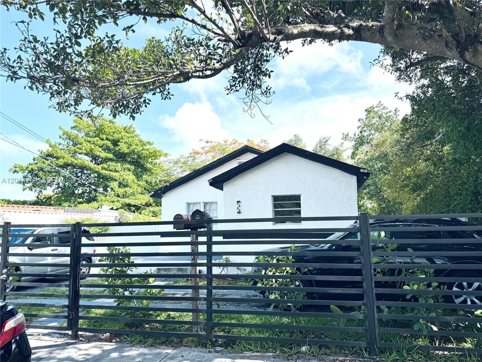 A white single-story house with a black roof is viewed behind a horizontal metal fence — 1484 NW 60th St, Miami FL 33142, MIAMI, FL