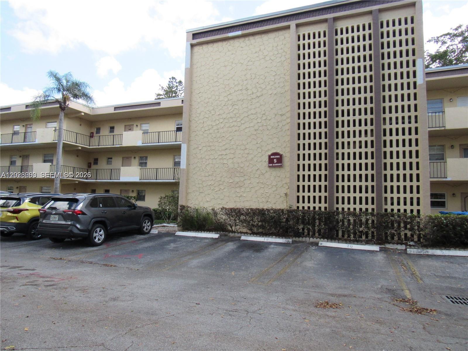 A three-story apartment building features a textured facade, decorative screen wall, and parking spaces — 100 Ashbury Rd # 104, Hollywood FL 33024, HOLLYWOOD, FL